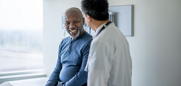 African senior man sits on exam table during a routine medical exam with his doctor standing to the side